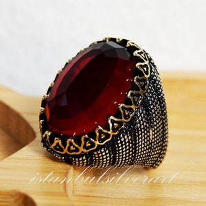 Puede incluir: Un anillo de plata con una gran piedra roja de forma ovalada engastada en un bisel dorado. La banda del anillo es texturizada y tiene un diseño decorativo.