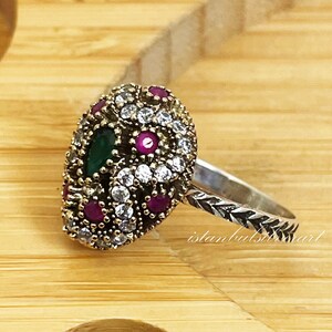 May include: A silver ring with a green and pink gemstone setting. The ring is decorated with small white stones and has a twisted band.