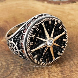 May include: A silver ring with a black compass face and a gold star design. The compass face has Roman numerals and red gemstones around the edge.