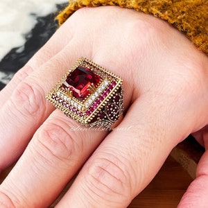 Peut inclure: Une bague en or avec une grande pierre rouge carrée entourée de petites pierres claires. La bague est sertie sur une bande en or.