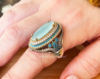 Puede incluir: Un anillo de plata con una gran piedra ovalada de color azul claro rodeada por una banda de piedras turquesas y blancas más pequeñas. El anillo está engastado en un metal dorado.