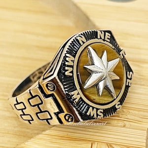 May include: A silver ring with a compass design. The compass face features a tiger's eye stone with a silver star in the center. The ring band has a watch-like design with a black and silver finish.