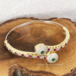 May include: A gold bangle bracelet with a delicate design featuring small red and white stones. The bracelet has two round charms with green and white stones. The bracelet is on a wooden surface.