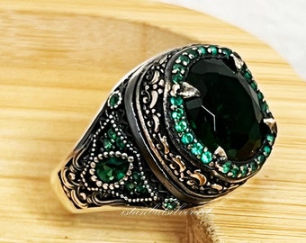 May include: A silver ring with an oval, dark green gemstone at the center, surrounded by smaller green stones. The ring has an ornate, detailed design with additional green accents. The ring is displayed on a light brown wooden surface.
