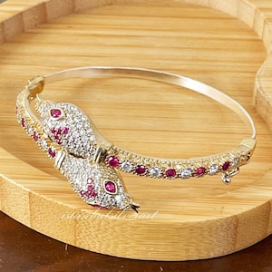 Può includere: Un braccialetto a forma di serpente in argento con pietre di zircone cubico rosse e trasparenti. Il braccialetto è su una superficie di legno.