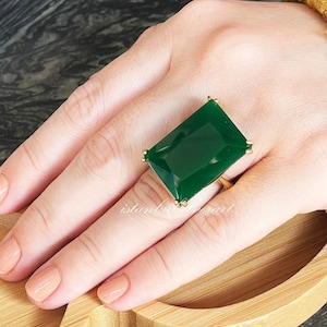 May include: A gold ring with a large, rectangular, emerald green gemstone. The ring is on a hand wearing a light brown nail polish.