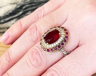 Könnte beinhalten: Ein goldener Ring mit einem großen ovalen roten Edelstein, umgeben von kleineren roten und weißen Edelsteinen.
