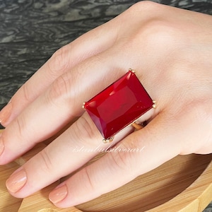 Könnte beinhalten: Ein goldener Ring mit einem großen, rechteckigen, roten Edelstein. Der Ring wird an einem Finger getragen.