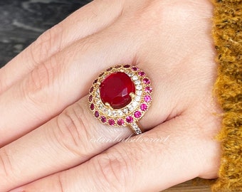 Könnte beinhalten: Ein goldener Ring mit einem großen roten ovalen Edelstein, der von kleineren roten und weißen Edelsteinen umgeben ist.