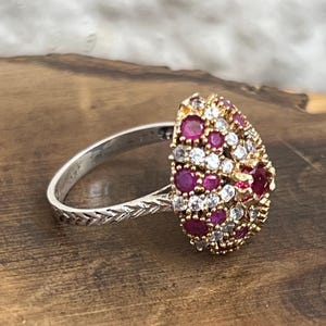 May include: A ring featuring a teardrop-shaped cluster head set with multiple round red stones and smaller clear white stones. The band is crafted from a silver-toned metal and displays a textured, braided-style pattern along the outer surface.