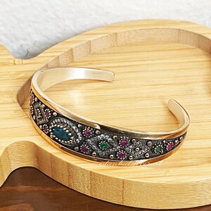 May include: A silver bangle bracelet with a gold-toned band and intricate design. The band is decorated with green and red gemstones and a larger blue gemstone in the center.