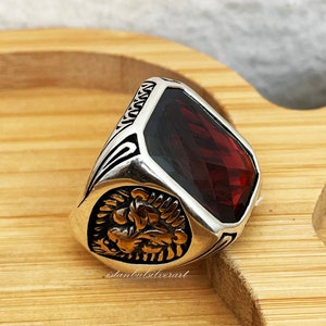 May include: A silver ring with a large, red, faceted gemstone. The ring has a detailed design with a gold lion head on the side.
