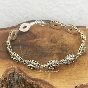 May include: A silver bracelet with a gold finish and red stones. The bracelet has a detailed design with small white stones around the red stones. The bracelet is on a wooden surface.