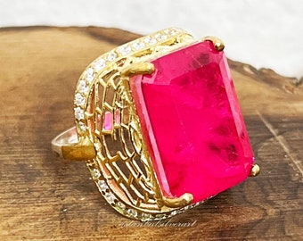 May include: A gold-toned ring featuring a large, rectangular, hot pink gemstone. The ring has a filigree design on the sides, accented with small, clear stones. The ring is set on a wooden surface.