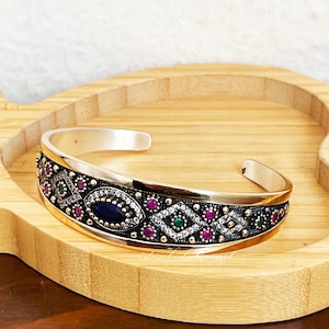 May include: A silver bangle bracelet with a gold-toned design featuring a blue oval stone surrounded by clear and pink stones. The bracelet has a geometric pattern with green and white accents.