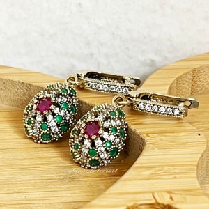 May include: A pair of dangle earrings featuring a red gemstone surrounded by clear and green stones. The earrings have a gold-tone finish and a hook fastening.