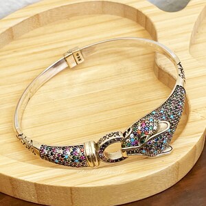May include: A silver bangle bracelet with a colorful, multi-faceted gemstone design. The bracelet has a unique clasp in the shape of a stylized animal head. The bracelet is marked "925" on the clasp.