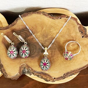 May include: A set of jewelry featuring a silver chain necklace, a pair of earrings, and a ring. Each piece has a teardrop-shaped pendant with a red and green gemstone design.