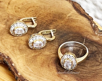 Könnte beinhalten: Ein Set aus goldfarbenem Schmuck mit einem Ring und einem Paar Ohrringen. Jedes Stück hat einen großen ovalen, klaren Stein, der von kleineren klaren Steinen umgeben ist.