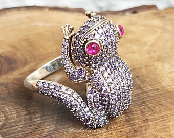 May include: A silver ring with a purple and pink frog design. The frog has pink eyes and is covered in small, sparkling stones.