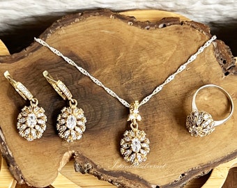 Könnte beinhalten: Ein Gold-Set mit Halskette, Ohrringen und Ring mit floralem Design und klaren Zirkonia-Steinen. Der Schmuck ist auf einer Holzoberfläche abgelegt.