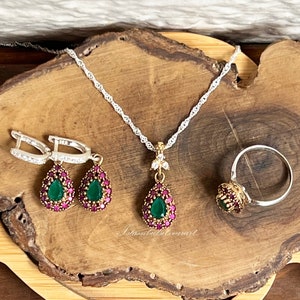 May include: A silver necklace, ring, and earrings set with green and pink gemstone accents. The jewelry is displayed on a wooden surface.