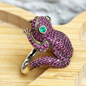 Puede incluir: Un anillo de plata con un diseño de rana. La rana está cubierta de piedras preciosas rosas y tiene dos piedras preciosas verdes para los ojos.