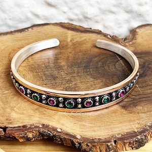 May include: A silver and rose gold bangle bracelet with a decorative band of green and pink gemstones.