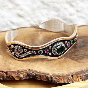May include: A silver and gold bangle bracelet with a black enamel design and green and red gemstones. The bracelet has a wavy shape and is decorated with intricate details.