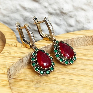 Puede incluir: Un par de pendientes colgantes con una piedra roja en forma de lágrima rodeada de piedras verdes y detalles plateados. Los pendientes tienen un aro plateado con piedras transparentes.