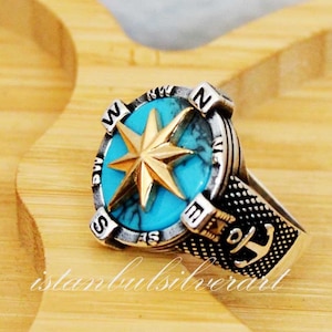 May include: A silver ring with a compass design featuring a turquoise stone and a gold star. The band of the ring has an anchor design.