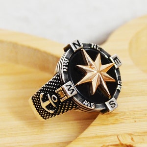 May include: A silver ring with a black compass face and a gold star. The ring also features an anchor on the side.