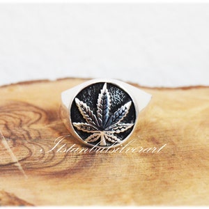 Puede incluir: Un anillo de plata con un dise&ntilde;o de hoja de marihuana negra. El anillo est&aacute; sobre una superficie de madera.