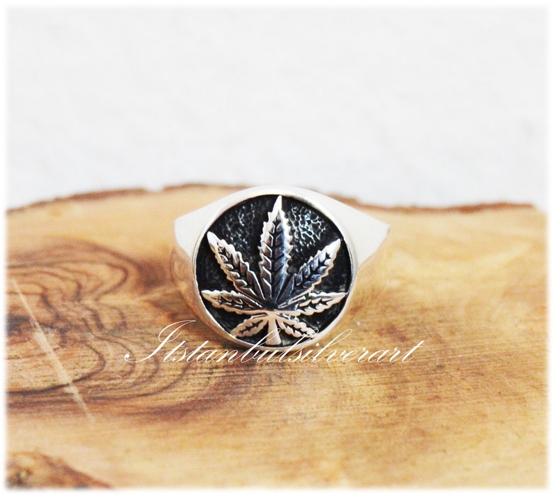 Puede incluir: Un anillo de plata con un dise&ntilde;o de hoja de marihuana negra. El anillo est&aacute; sobre una superficie de madera.