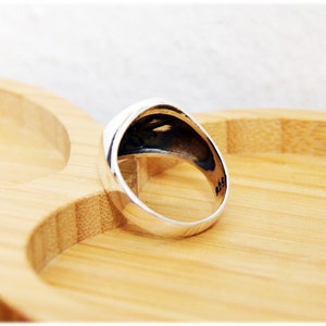 Puede incluir: Un anillo de plata con la parte superior redondeada y un acabado liso y pulido. El anillo est&aacute; sobre una superficie de madera.