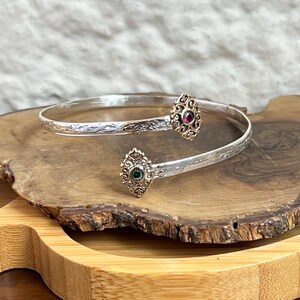 Puede incluir: Un brazalete plateado con detalles dorados. El brazalete presenta dos elementos decorativos ovalados, uno con una piedra roja y otro con una piedra verde. El brazalete se muestra sobre una superficie de madera.