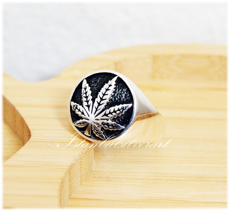 Puede incluir: Un anillo de plata con un dise&ntilde;o de hoja de marihuana en blanco y negro. El anillo est&aacute; sobre una superficie de madera.
