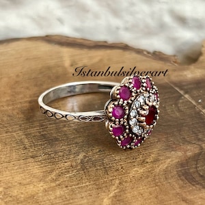 Könnte beinhalten: Ein silberner Ring mit einem floralen Design, das einen großen roten Edelstein zeigt, der von kleineren roten Edelsteinen und weißen Zirkonia-Steinen umgeben ist.