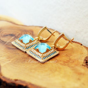 Puede incluir: Un par de pendientes de oro con piedras cuadradas azules engastadas en un marco de oro. Los pendientes tienen un dise&ntilde;o delicado con peque&ntilde;as piedras transparentes que rodean las piedras azules. Los pendientes est&aacute;n sobre una superficie de madera.