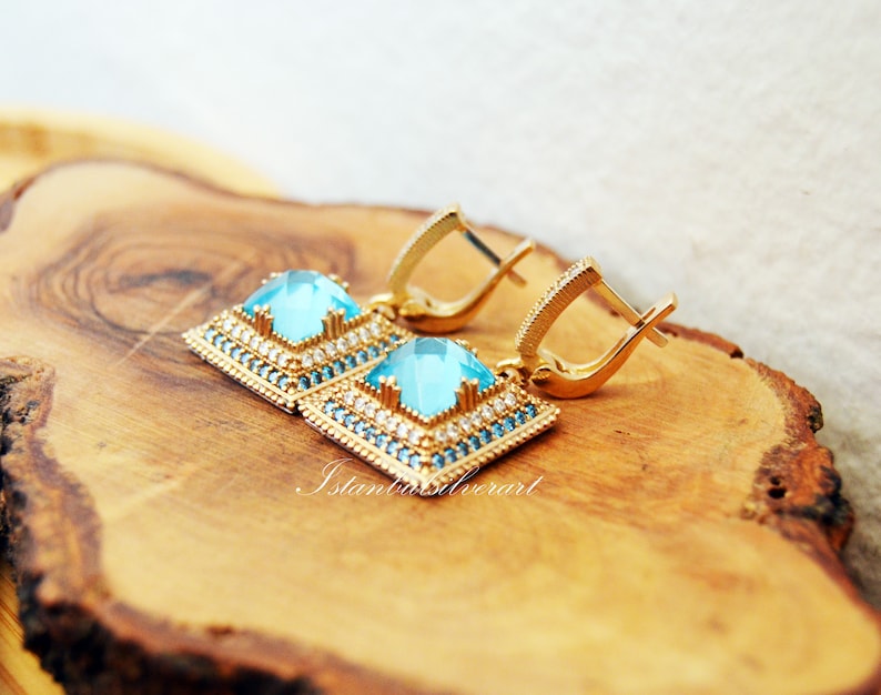 Puede incluir: Un par de pendientes de oro con piedras cuadradas azules engastadas en un marco de oro. Los pendientes tienen un dise&ntilde;o delicado con peque&ntilde;as piedras transparentes que rodean las piedras azules. Los pendientes est&aacute;n sobre una superficie de madera.