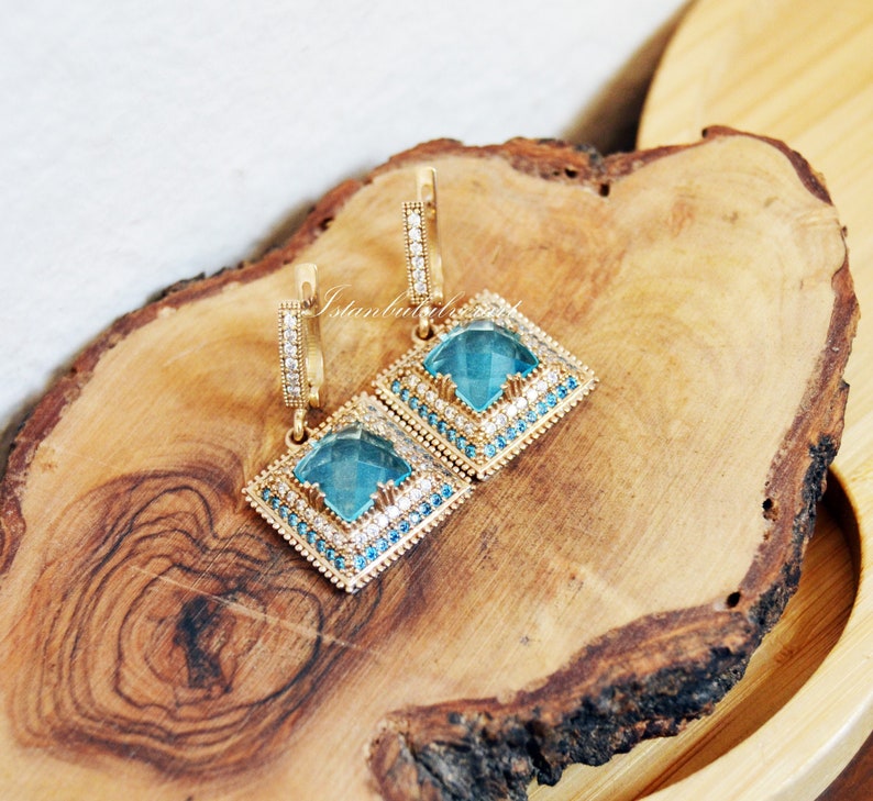 Puede incluir: Un par de pendientes de oro con detalles de piedras preciosas azules y forma cuadrada. Los pendientes tienen un dise&ntilde;o colgante con un peque&ntilde;o aro en la parte superior.