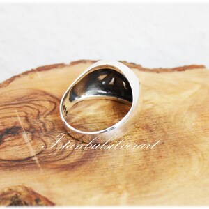 Puede incluir: Un anillo de plata con un dise&ntilde;o en relieve en la parte superior. El anillo est&aacute; sobre una superficie de madera.