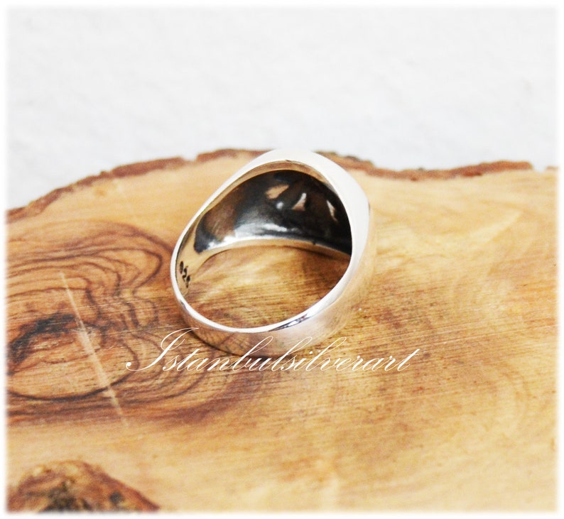 Puede incluir: Un anillo de plata con un dise&ntilde;o en relieve en la parte superior. El anillo est&aacute; sobre una superficie de madera.