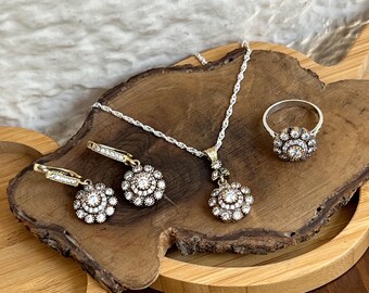 Könnte beinhalten: Eine silberne Kettenkette, ein Ring und ein Paar Ohrringe mit einem floralen Design und klaren Steinen. Der Schmuck wird auf einer Holzoberfläche präsentiert.