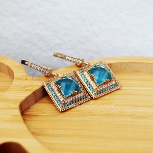 Puede incluir: Un par de pendientes de plata con una piedra preciosa azul cuadrada rodeada de piedras preciosas transparentes m&aacute;s peque&ntilde;as. Los pendientes tienen un dise&ntilde;o colgante y est&aacute;n sobre una superficie de madera.