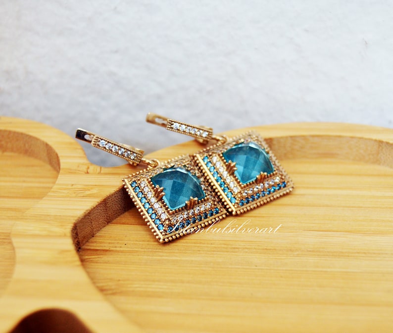 Puede incluir: Un par de pendientes de plata con una piedra preciosa azul cuadrada rodeada de piedras preciosas transparentes m&aacute;s peque&ntilde;as. Los pendientes tienen un dise&ntilde;o colgante y est&aacute;n sobre una superficie de madera.