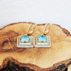 Puede incluir: Un par de pendientes de oro con una piedra preciosa azul cuadrada rodeada de piedras preciosas m&aacute;s peque&ntilde;as transparentes y azules. Los pendientes est&aacute;n sobre una superficie de madera.