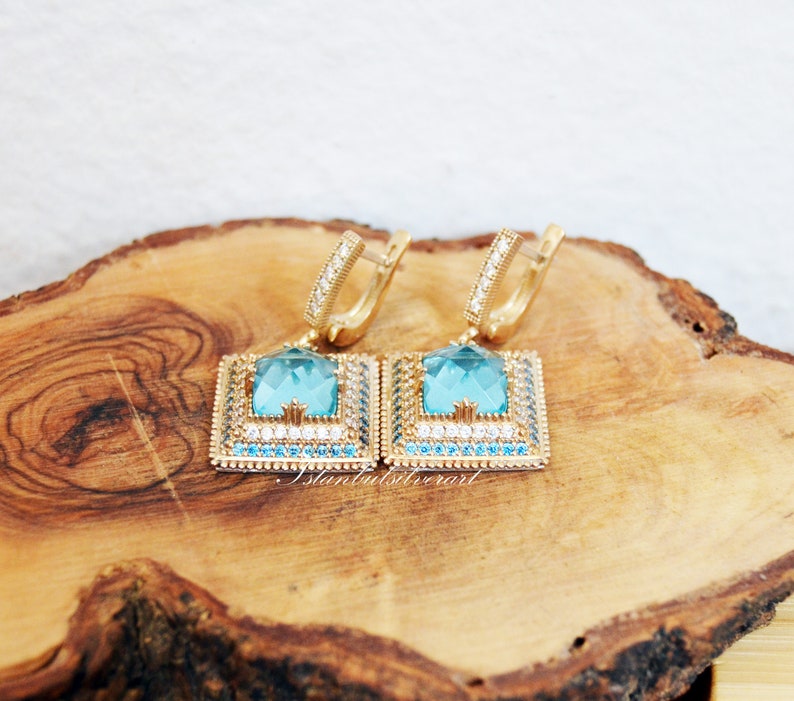 Puede incluir: Un par de pendientes de oro con una piedra preciosa azul cuadrada rodeada de piedras preciosas m&aacute;s peque&ntilde;as transparentes y azules. Los pendientes est&aacute;n sobre una superficie de madera.