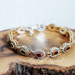 Puede incluir: Un brazalete dorado con piedras rojas y transparentes. El brazalete está hecho de plata y tiene un diseño delicado.