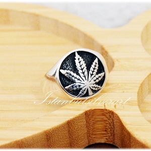 Puede incluir: Un anillo de plata con un dise&ntilde;o de hoja de marihuana negra. El anillo est&aacute; sobre una superficie de madera.